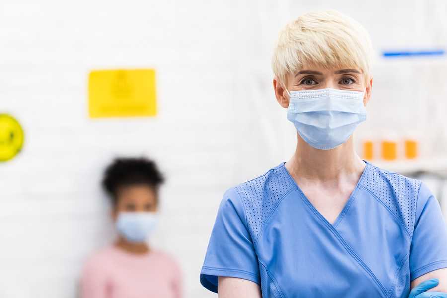A doctor wearing a face mask, standing with her arms crossed in a vaccination clinic, with a patient blurred in the background