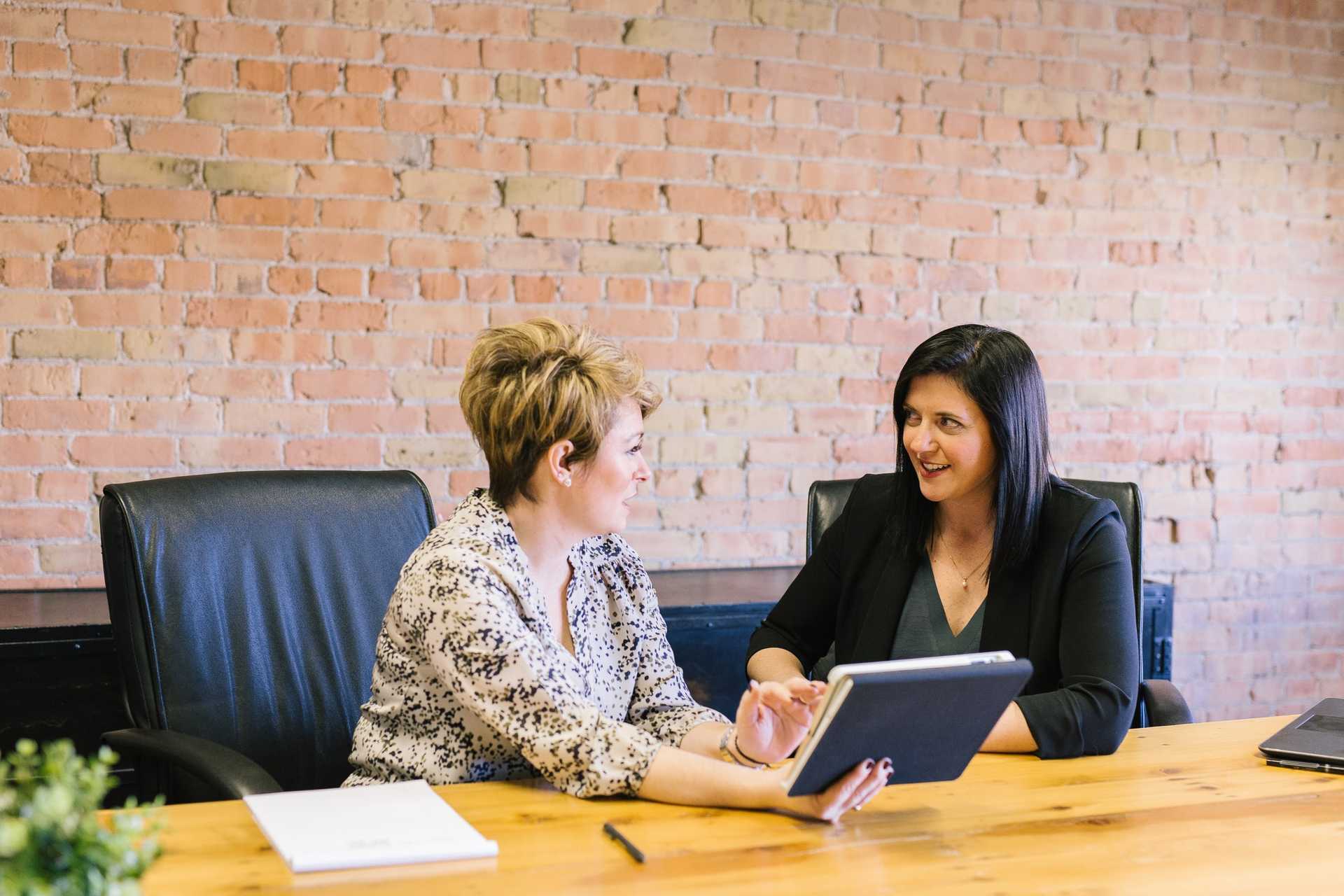 Two women sitting on leather office chairs, one showing the other something on her notebook