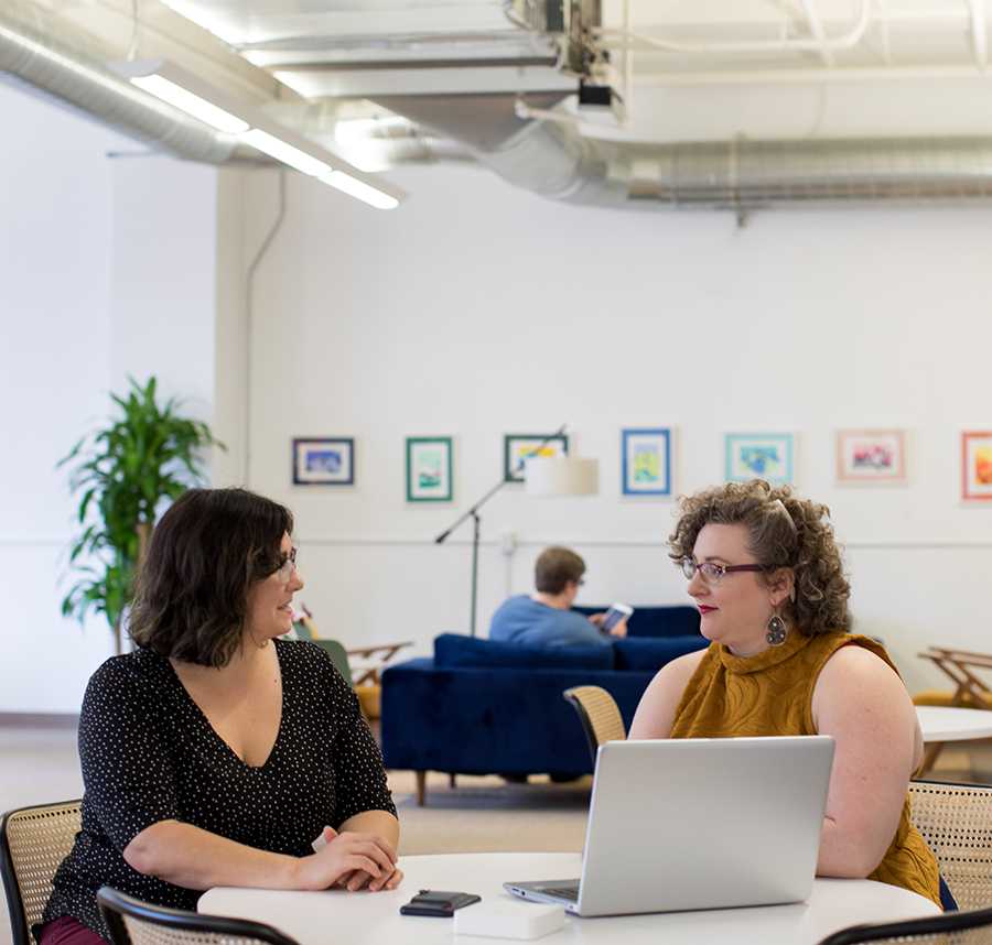Two women talk to each other across a round desk. They are both dressed professionally.