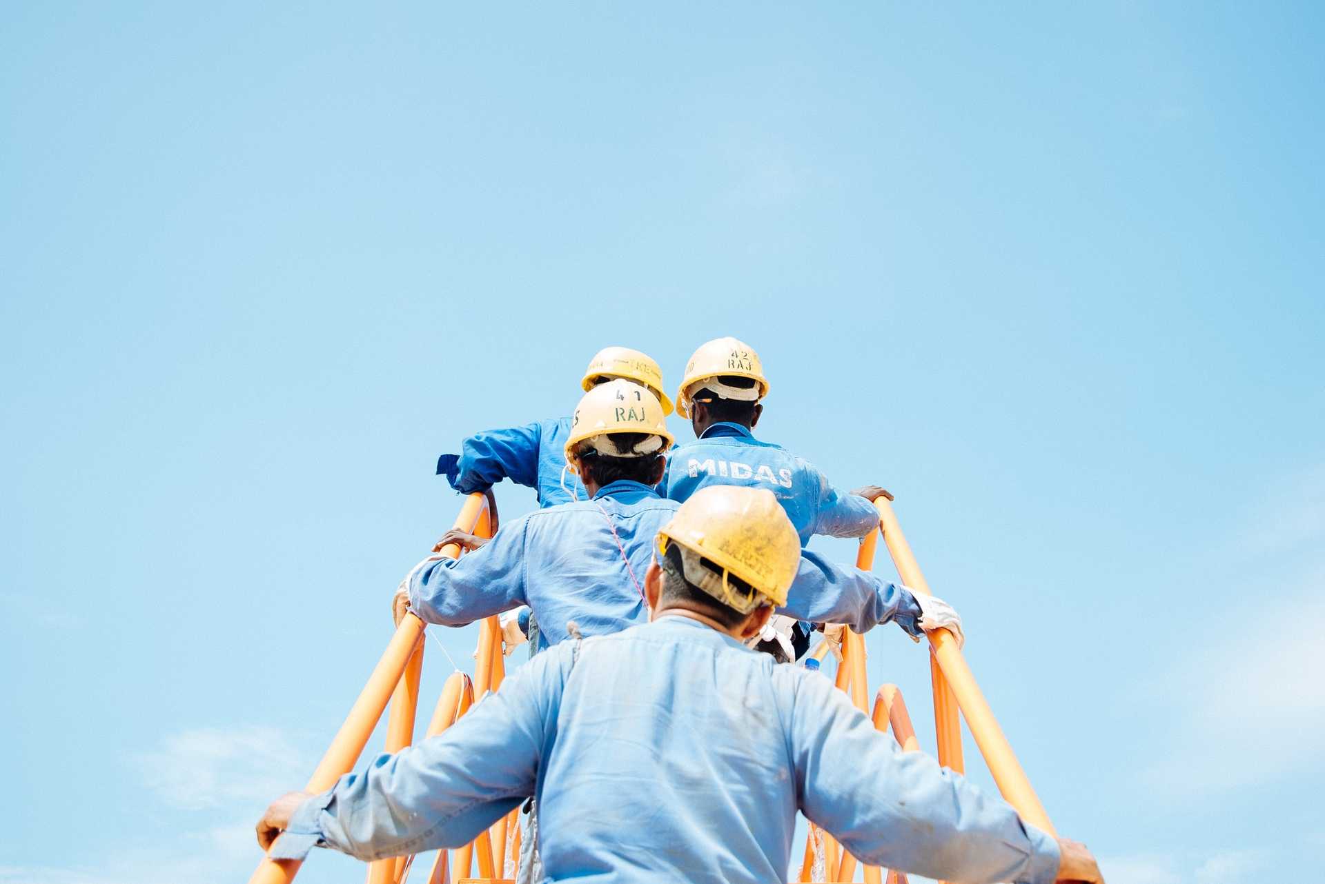 Four individuals dressed in blue shirts and wearing hards climb a railing, with their backs facing the camera