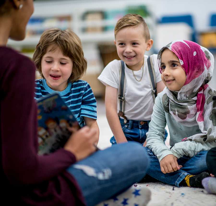 A multicultural group of children sit on the floor. Their teacher sits across from them reading them a book.