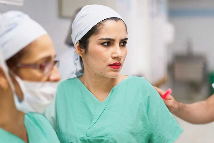 A hospital worker looks worried and uncomfortable as someone touches her shoulder from behind her.