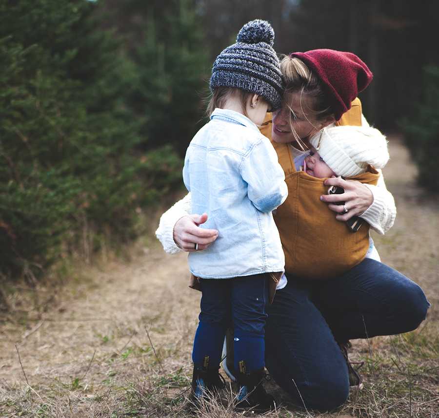 A mother kneels down to comfort her young daughter. The mother carries another child in a baby carrier across her front.