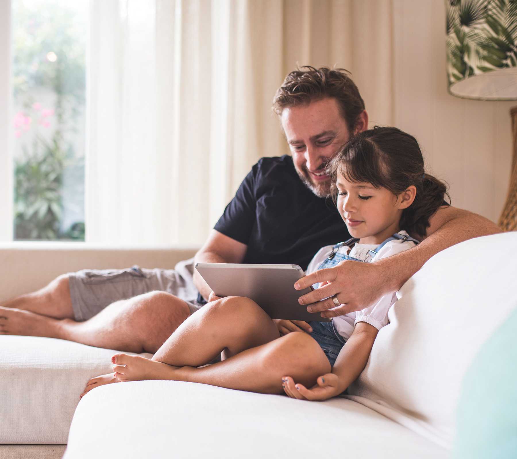 Father and daughter sitting on a sofa. Dad is holding a digital tablet and they are both looking at it..